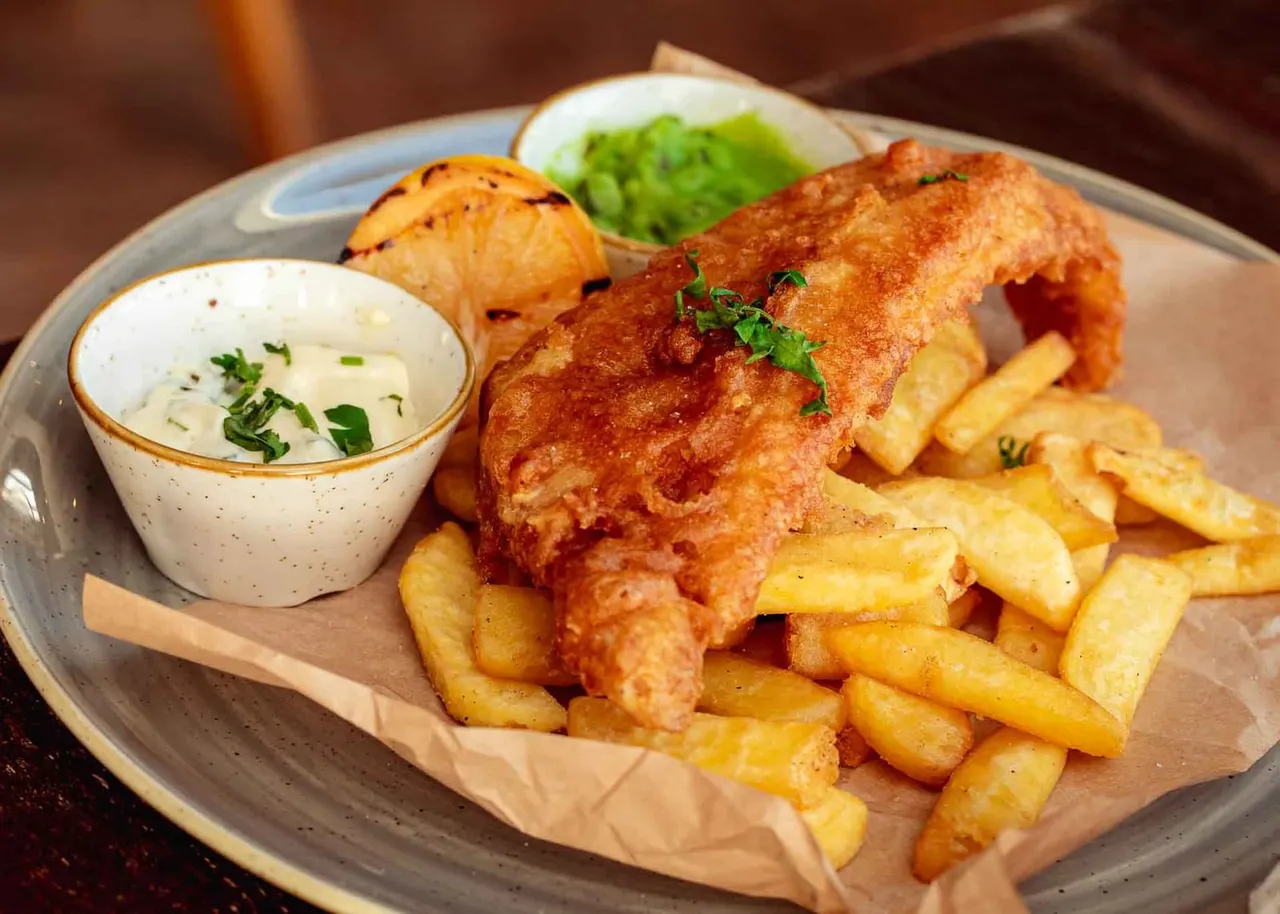 Golden-battered fish fillet with chips, tartare sauce, and coleslaw on a wooden board.