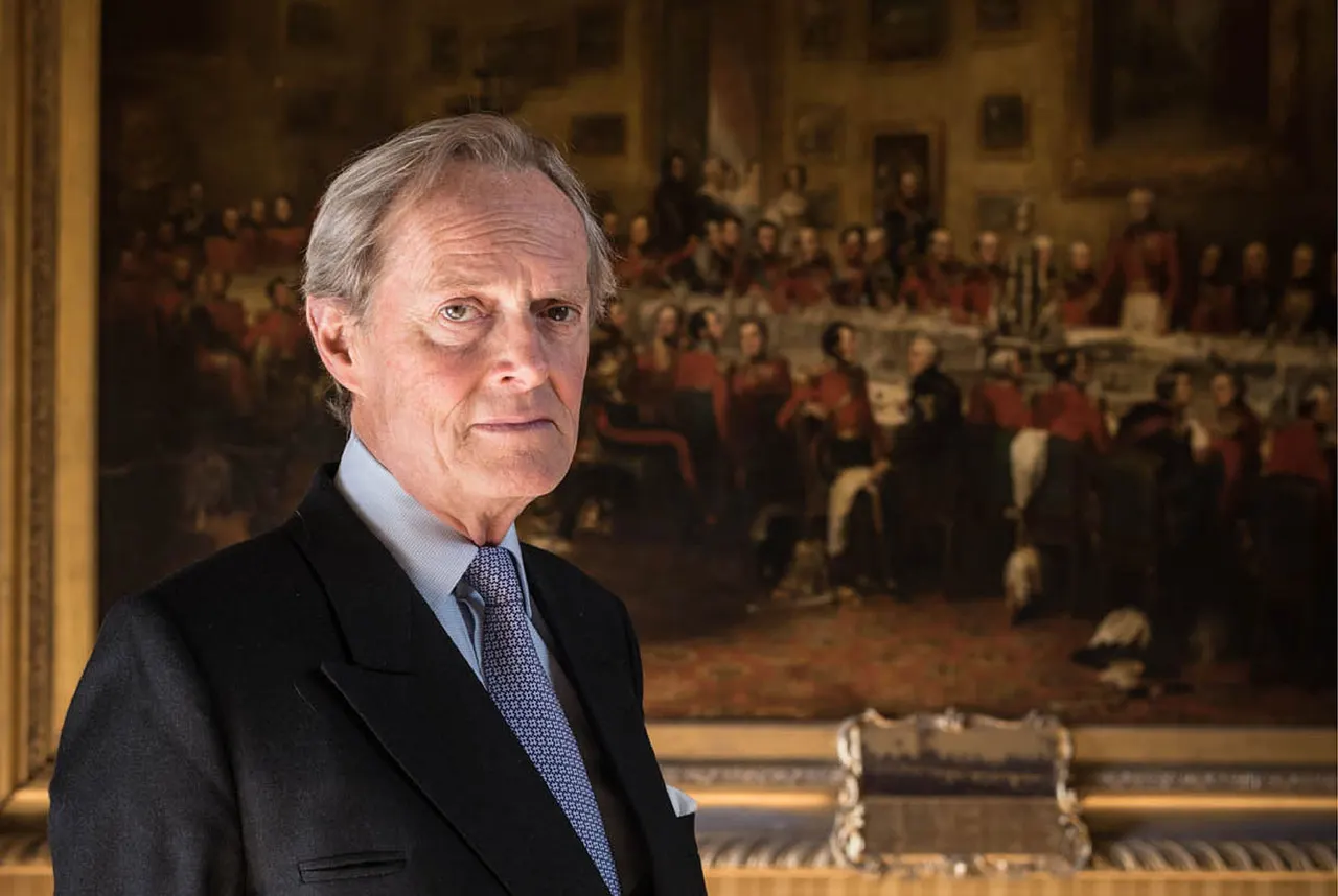 Older man in dark suit and blue tie standing in front of a large historical painting depicting a crowded formal gathering in an ornate interior.