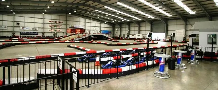 Indoor go-kart racing track with multiple lanes, barriers and spectator areas in a large warehouse facility.
