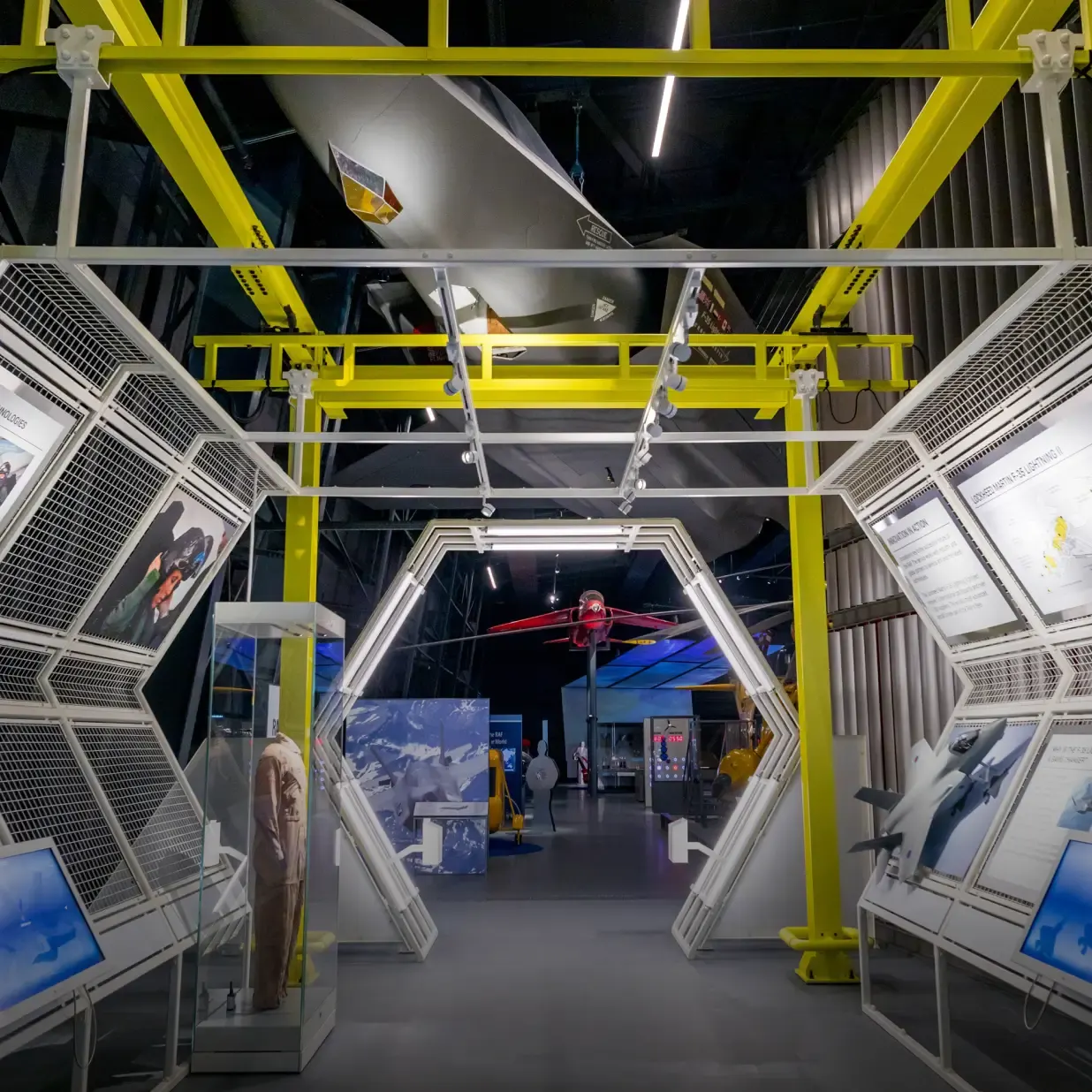 Interior of aviation museum gallery with yellow structural framework, display panels, and red aircraft suspended from ceiling.