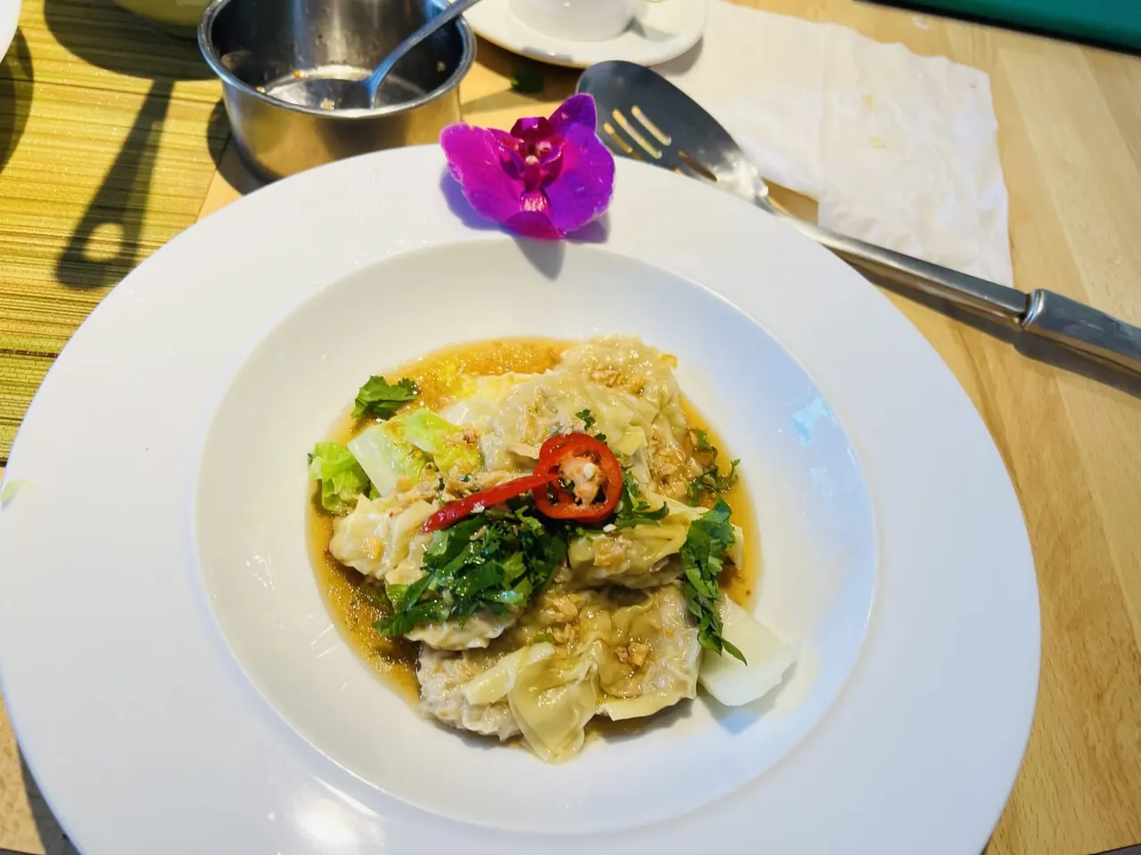 A plate of steamed dumplings garnished with fresh coriander, chilli slices and a purple orchid flower, served at a dining table.
