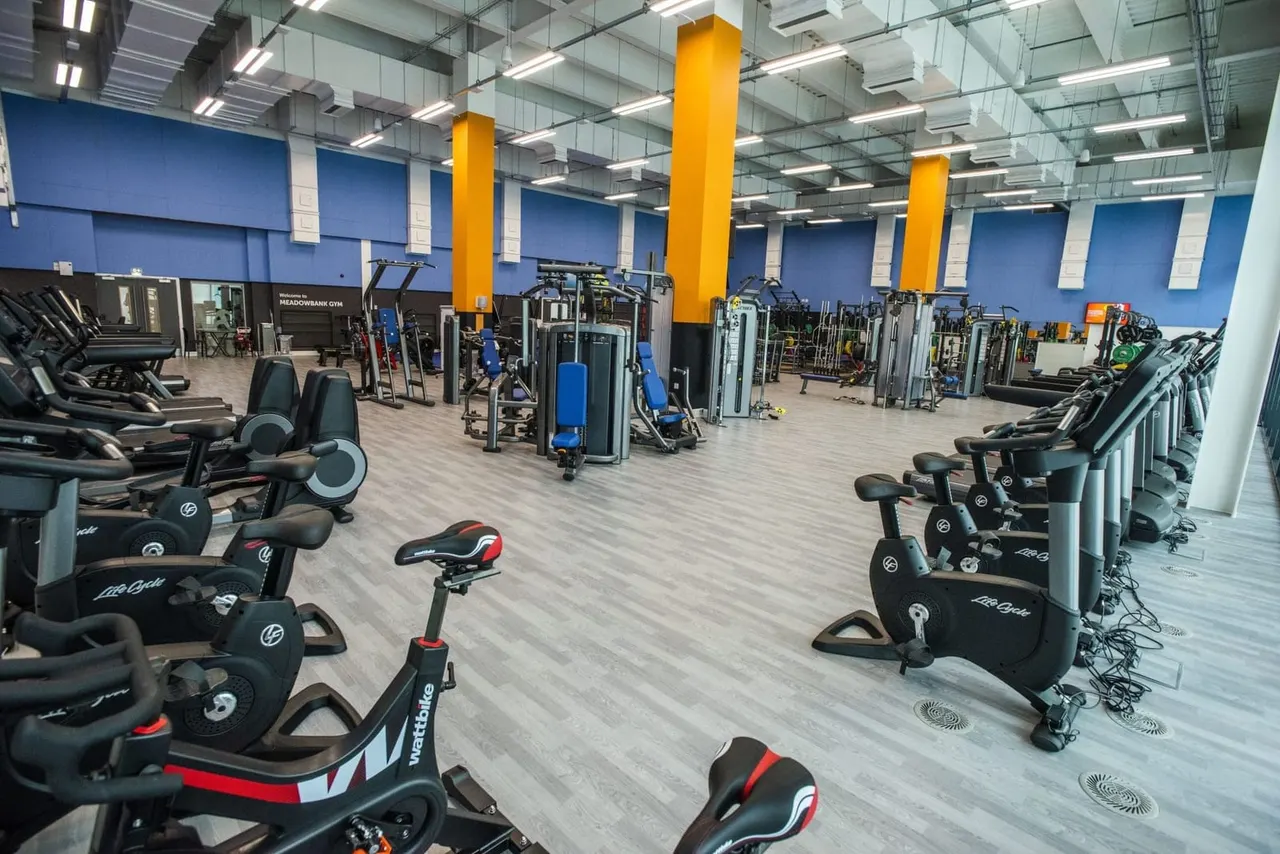 Spacious gym interior with rows of exercise bikes and cardio machines on either side, strength training equipment in the centre, and bright yellow support pillars.