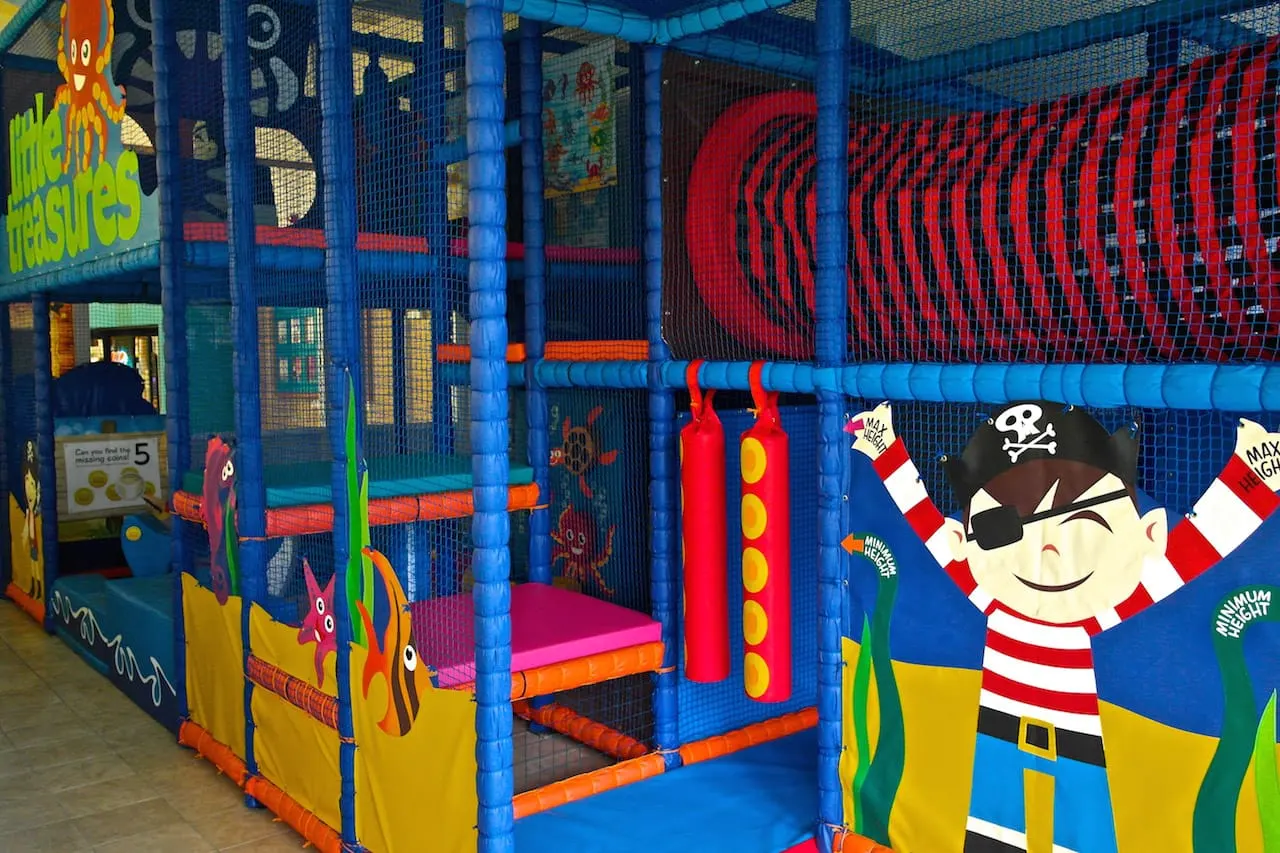 Colourful indoor soft play structure with blue frame, red and yellow padded climbing elements, and pirate-themed artwork on panels.