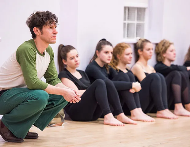 Male instructor crouching beside a line of seated young people during a dance or movement class.