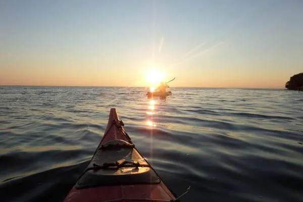 Kayaker paddling on calm water at sunset, viewed from inside the kayak looking towards the golden sun on the horizon.