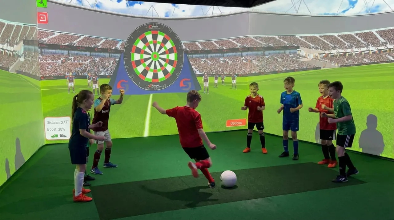 Children playing football in an indoor virtual reality stadium with a large dartboard target displayed above.