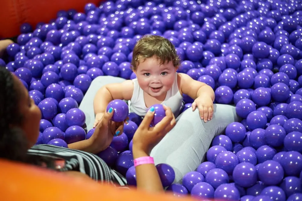 A toddler in a white vest plays in a ball pit filled with purple balls, holding two balls while smiling at an adult.