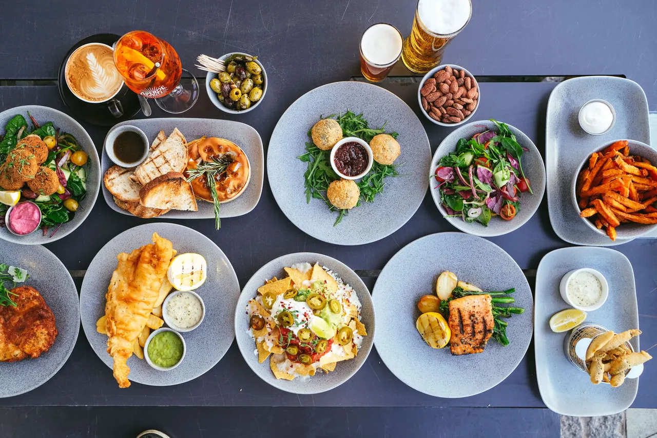 Overhead view of plated dishes including fish, salads, fried items and breads, with beers and cocktails arranged on a dark table.