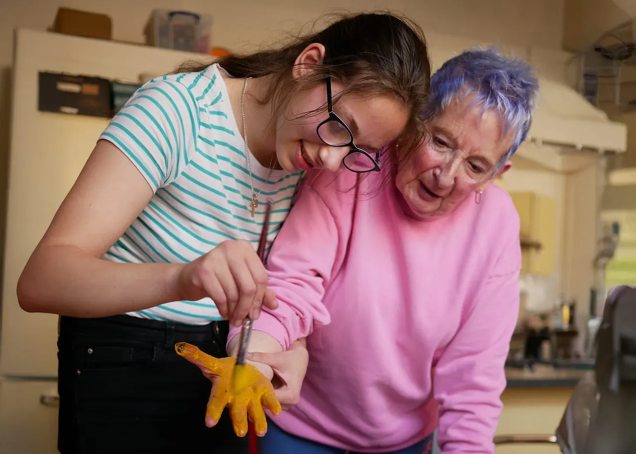 A young woman and older adult smiling together whilst holding a yellow toy chicken in an indoor activity space.