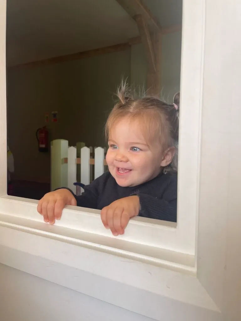A toddler smiles whilst peering through a white-framed window inside an indoor play centre.