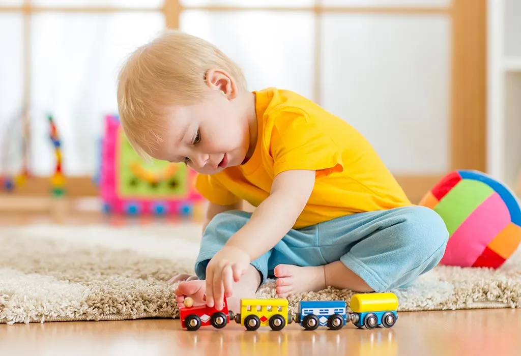 Toddler in yellow shirt playing with toy trains on a carpet in a bright playroom.
