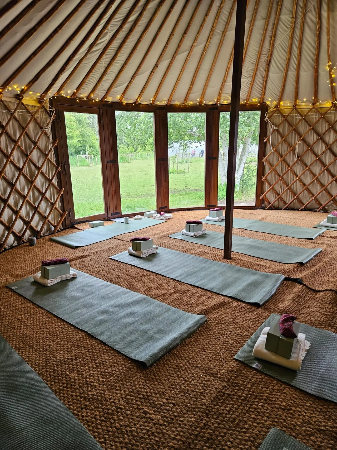 Interior of a yurt with yoga mats laid out on the floor, overlooking green fields through large windows.
