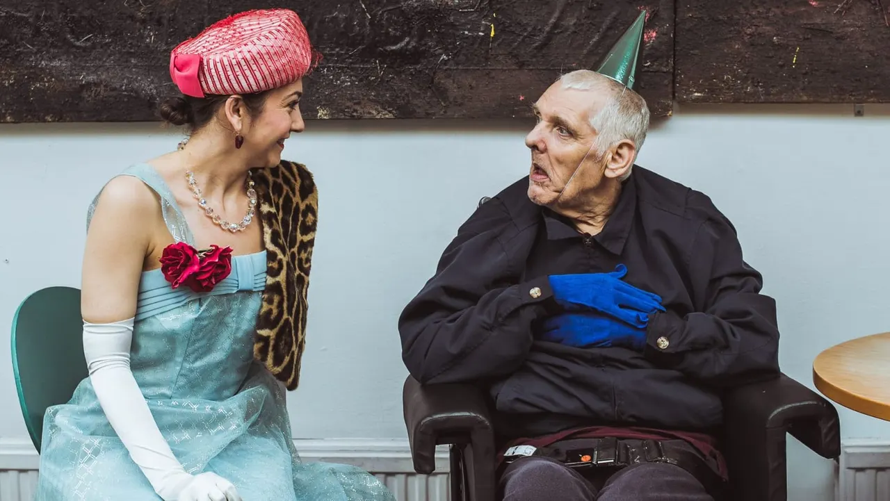 Two people in party hats and festive outfits sit facing each other on chairs, appearing to have a lively conversation.