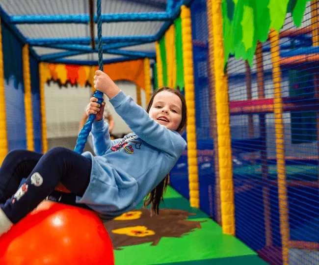 Young child playing on a red bouncy ball inside a colourful indoor soft play area with netting and foam structures.