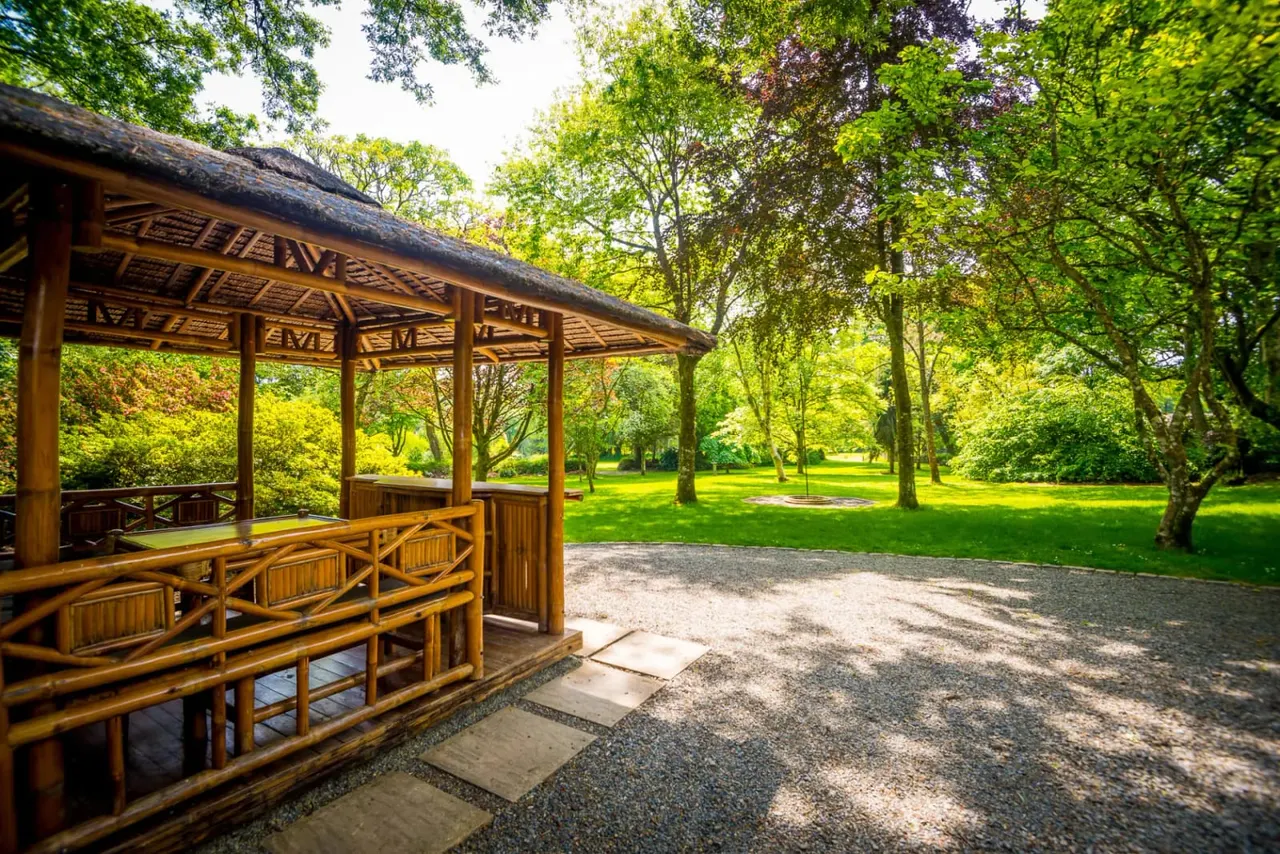 Wooden pavilion with thatched roof beside a gravel path in a sunlit park surrounded by mature trees and green lawns.