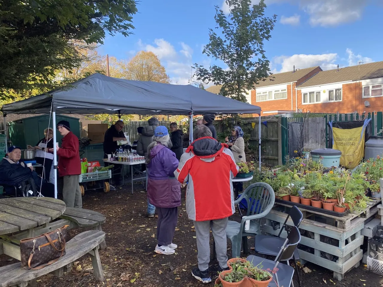 Community garden gathering with visitors under a gazebo tent, surrounded by raised vegetable beds and potted plants, with residential buildings visible beyond.