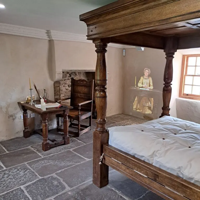 Historic bedroom interior with wooden four-poster bed, writing desk, chair and period furnishings in a stone-flagged room.