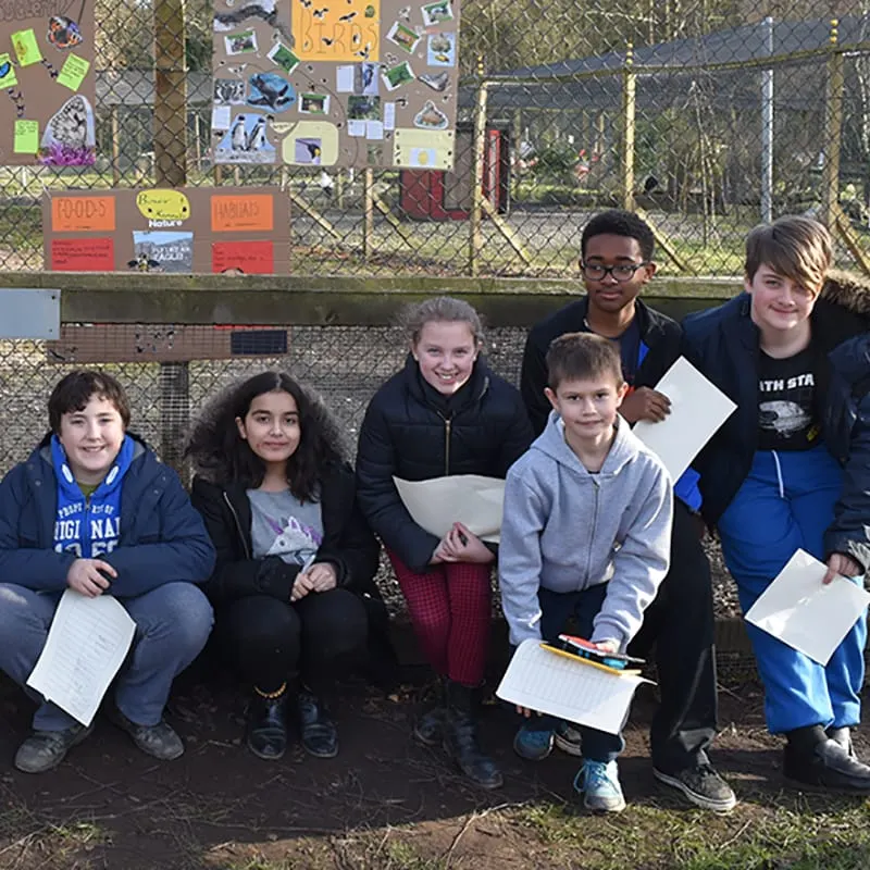 Group of children holding clipboards and papers posing together outdoors against a chain-link fence with animal enclosures visible behind them.