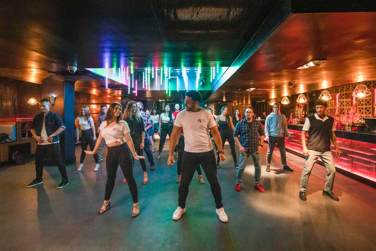 Group of people dancing together in a neon-lit venue with colourful overhead lighting and red bar seating.