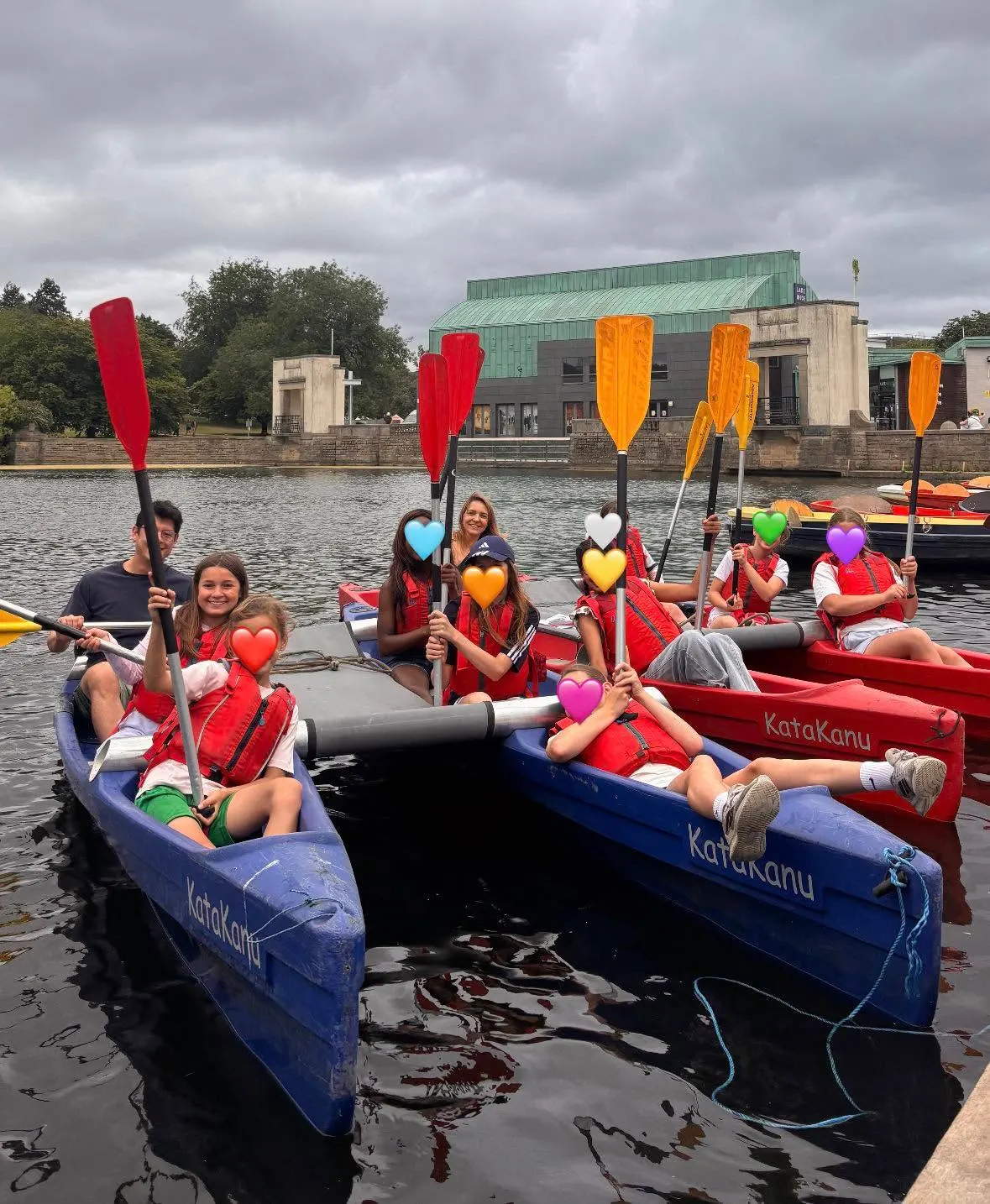 Groups of people kayaking on a river, wearing red life jackets and holding colourful paddles with heart-shaped decorations.