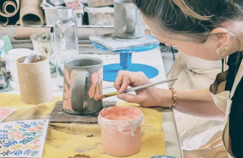 Woman hand-painting a ceramic vessel with a brush at a pottery workbench.