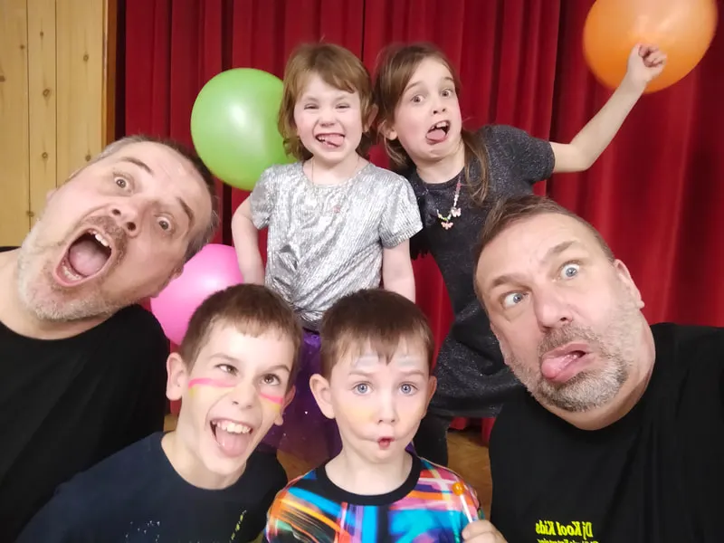 Two adults and four children pulling silly faces at a party with red curtains and coloured balloons behind them.