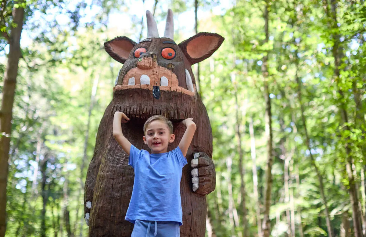A young child in a blue shirt posing with arms raised inside a large wooden creature sculpture in a woodland setting.