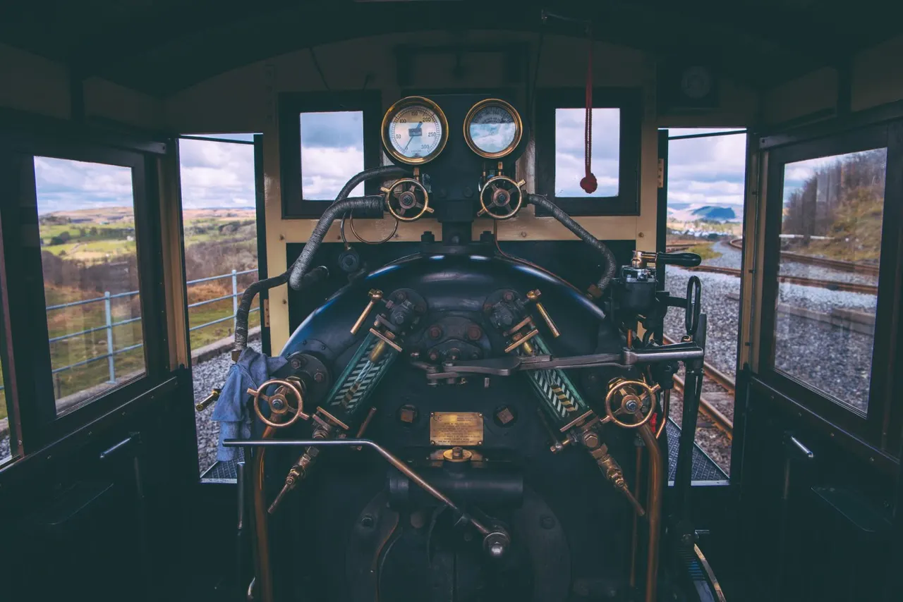 Interior of a vintage steam locomotive cab with steering wheel, gauges and controls, viewed through windows overlooking rural countryside and railway tracks.