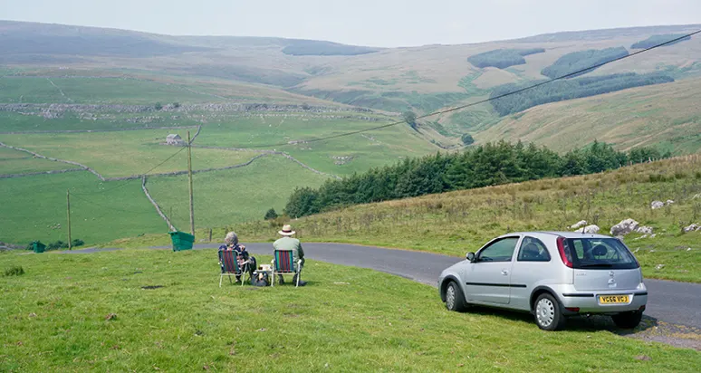 Two people sit in camping chairs on a grassy hillside beside a parked car, overlooking rolling valleys and farmland.