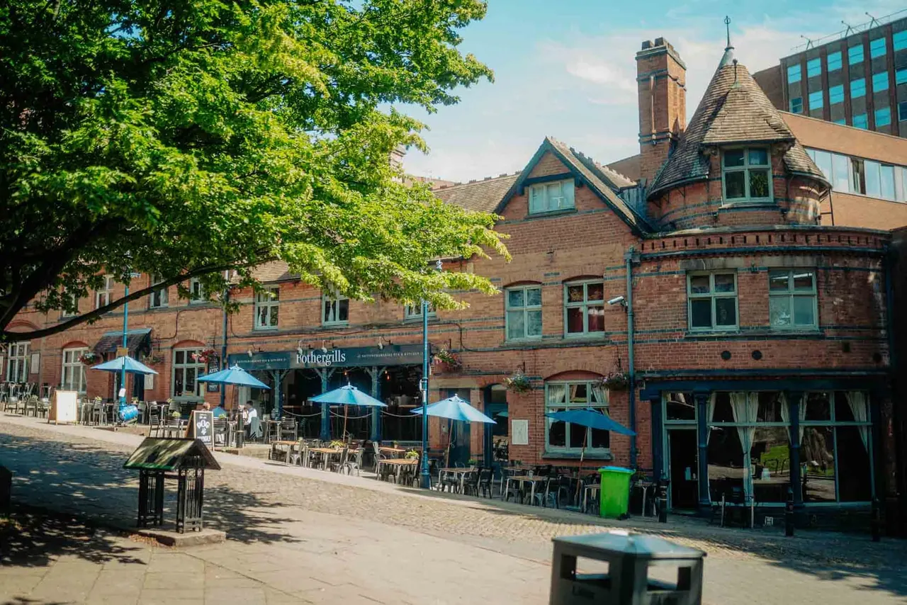 Historic brick building with outdoor seating under blue umbrellas, tree-lined courtyard, and diners enjoying the sunny street-facing patio.