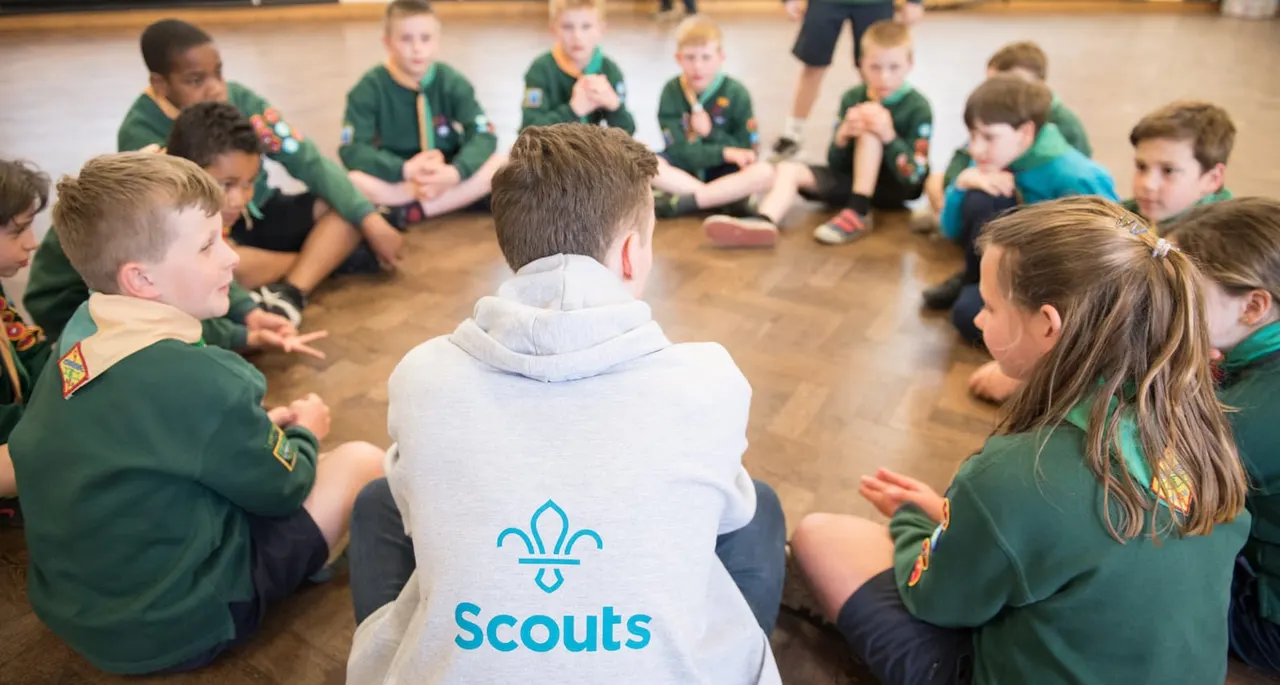 A scout leader wearing a Scouts gilet sits on a wooden floor surrounded by children in green uniforms during an indoor activity session.