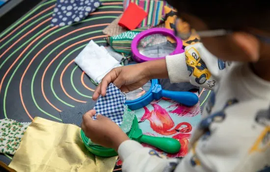Child's hands holding colourful sensory toys and textured fabric samples on a patterned mat.