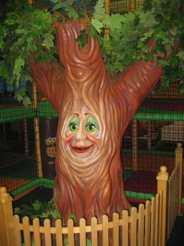 Carved wooden tree sculpture with a smiling face inside a soft play area, surrounded by green foliage and a yellow fence.