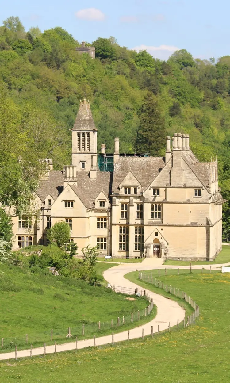 Victorian mansion with tower and chimneys set in wooded hillside with manicured grounds and winding pathway.