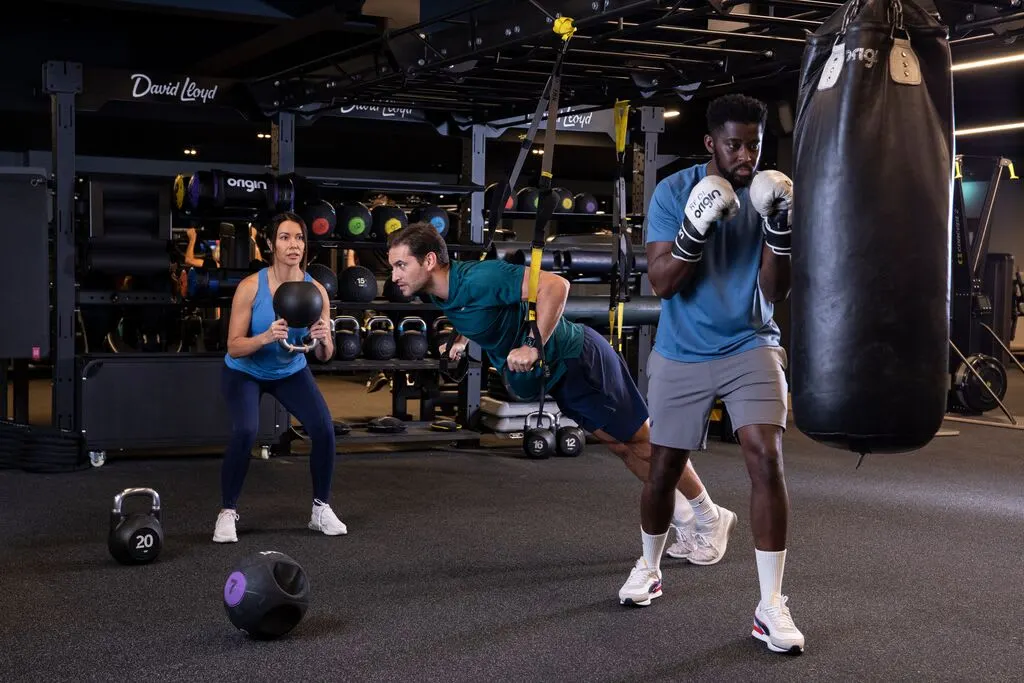 Three people exercise in a gym, with a man boxing a heavy bag whilst two others hold kettlebells and a medicine ball nearby.