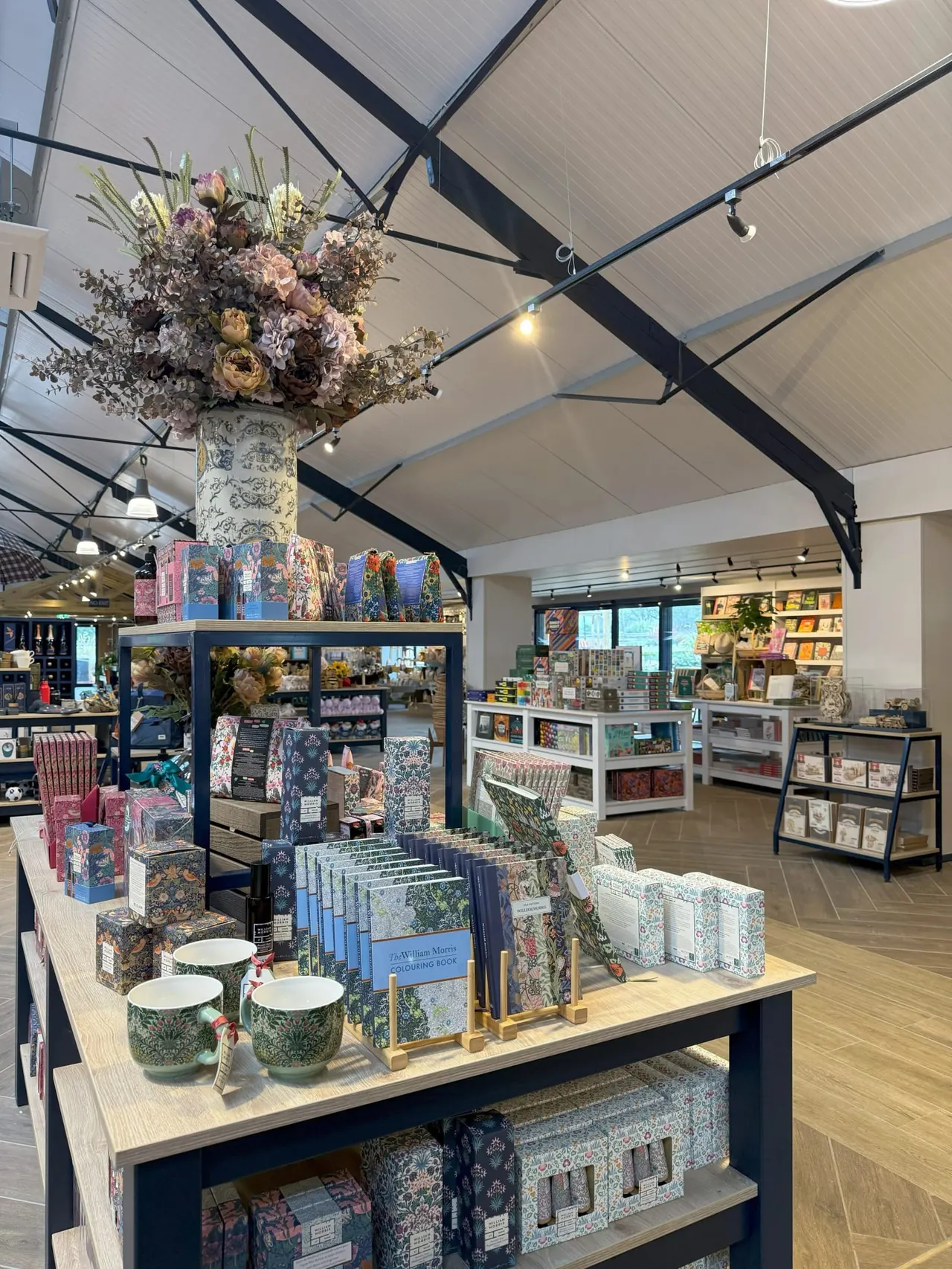 Interior of a gift shop with display tables featuring books, decorative items and patterned homeware, beneath a large floral arrangement hanging from the ceiling.