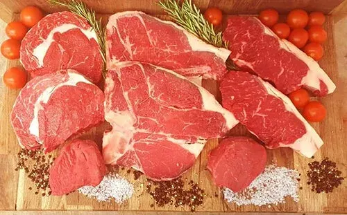 Raw red meat cuts arranged on a wooden board with fresh tomatoes, rosemary and spices.