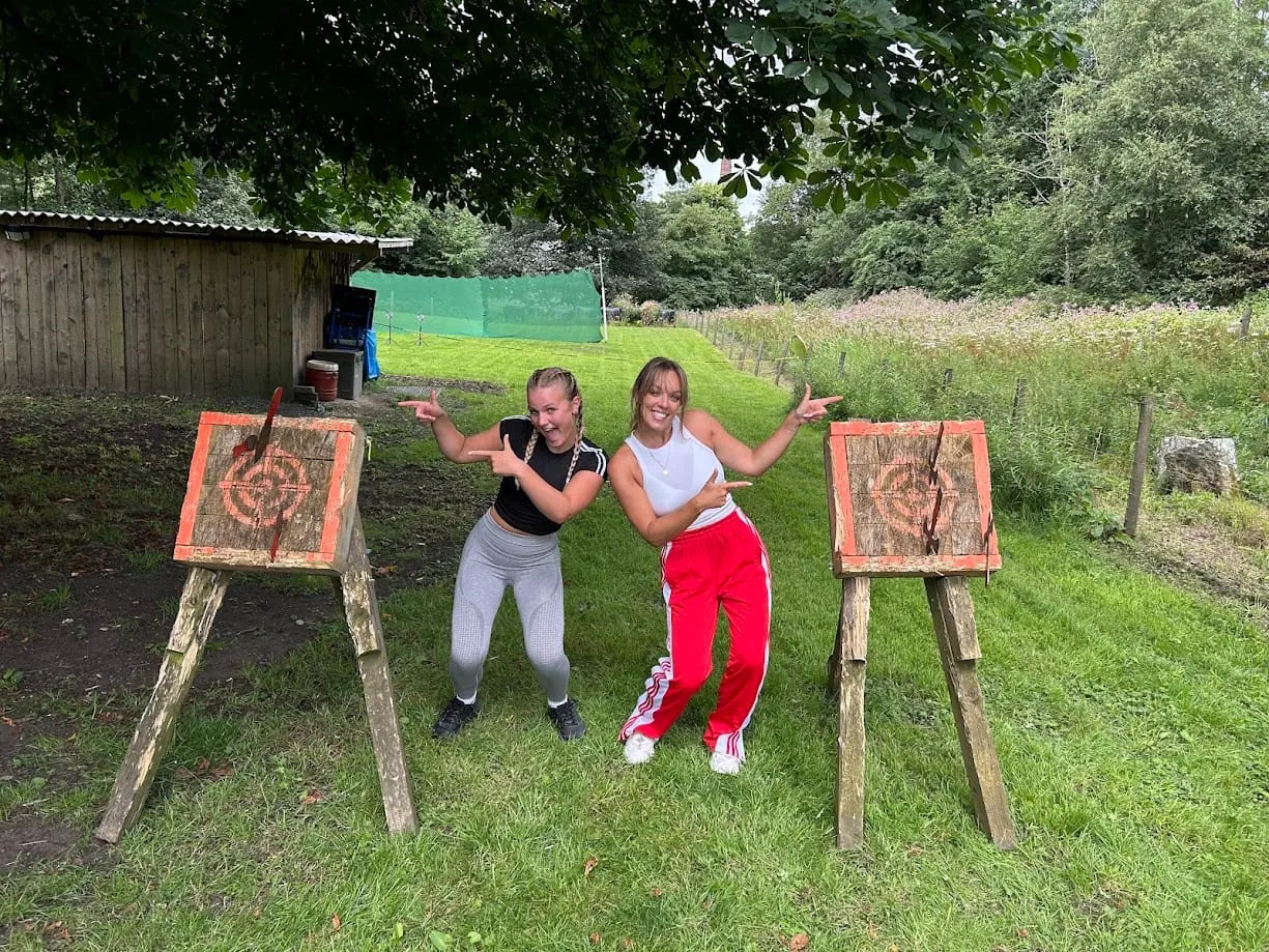 Two young women posing between axe-throwing targets in a grassy outdoor activity area surrounded by trees.