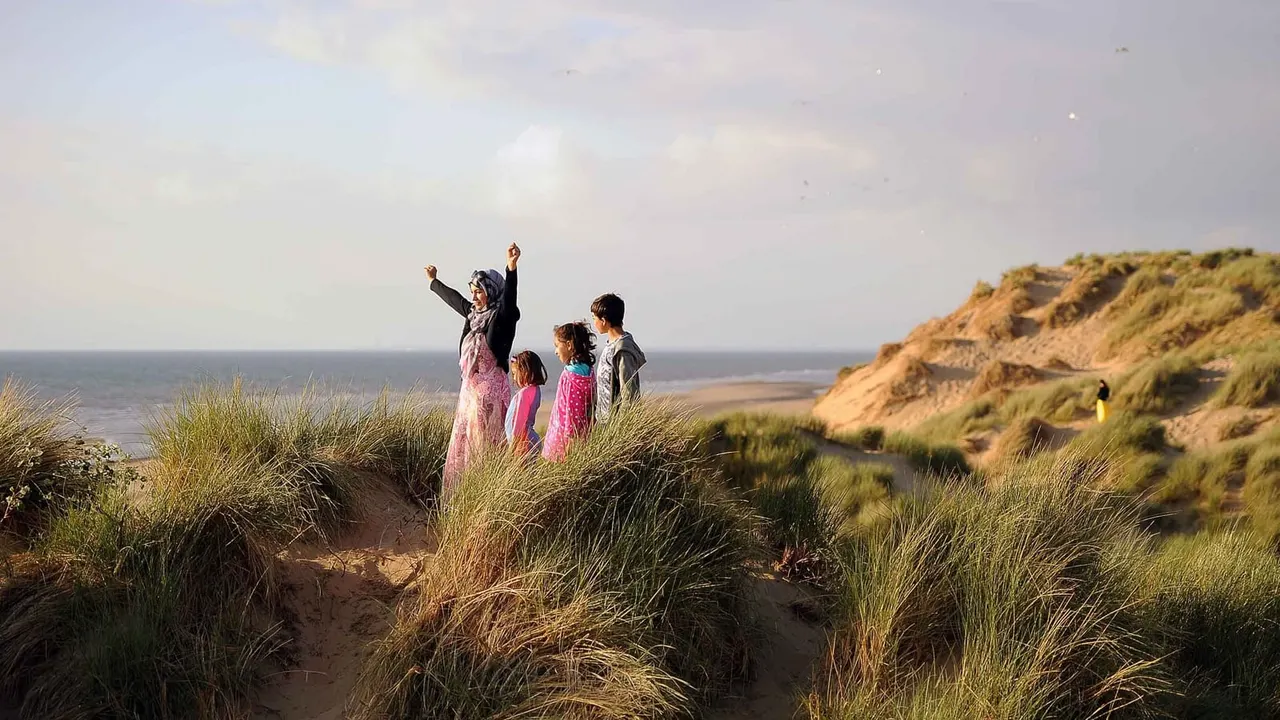Adult and children standing on sand dunes with marram grass, facing the sea and sandy cliffs on a clear day.
