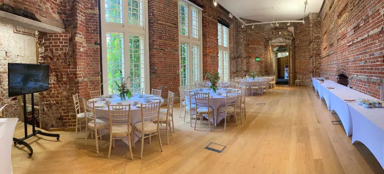 Event space with round tables set for dining, wooden chairs, white tablecloths and exposed red brick walls with large windows.
