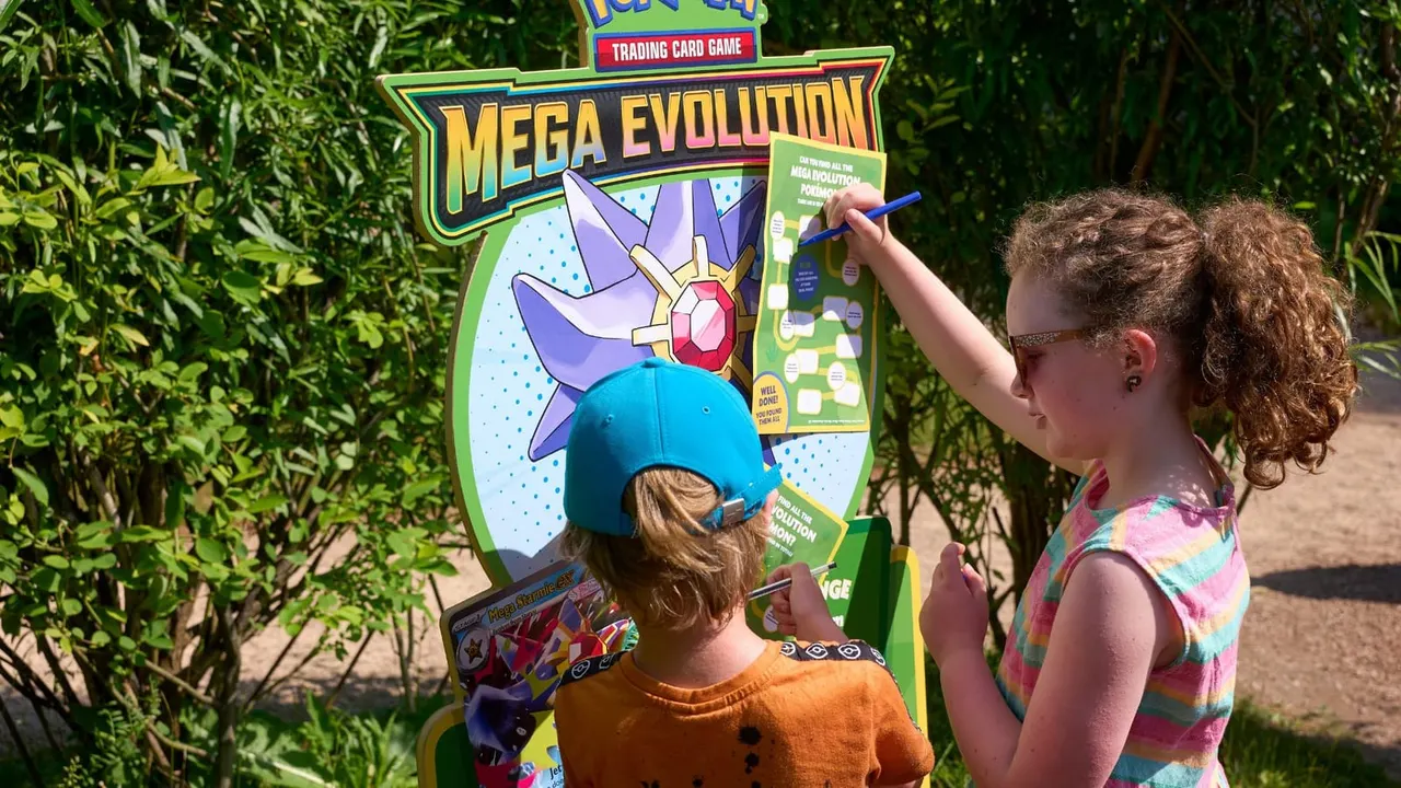 Two children playing an interactive Pokémon Mega Evolution trading card game display outdoors surrounded by green foliage.