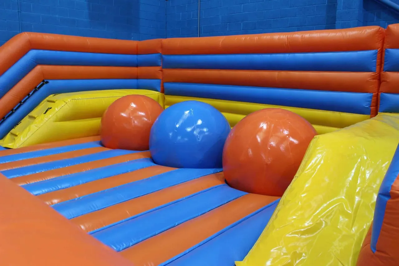 Inflatable obstacle course with alternating blue and orange padded sections, yellow foam barriers and large coloured balls.