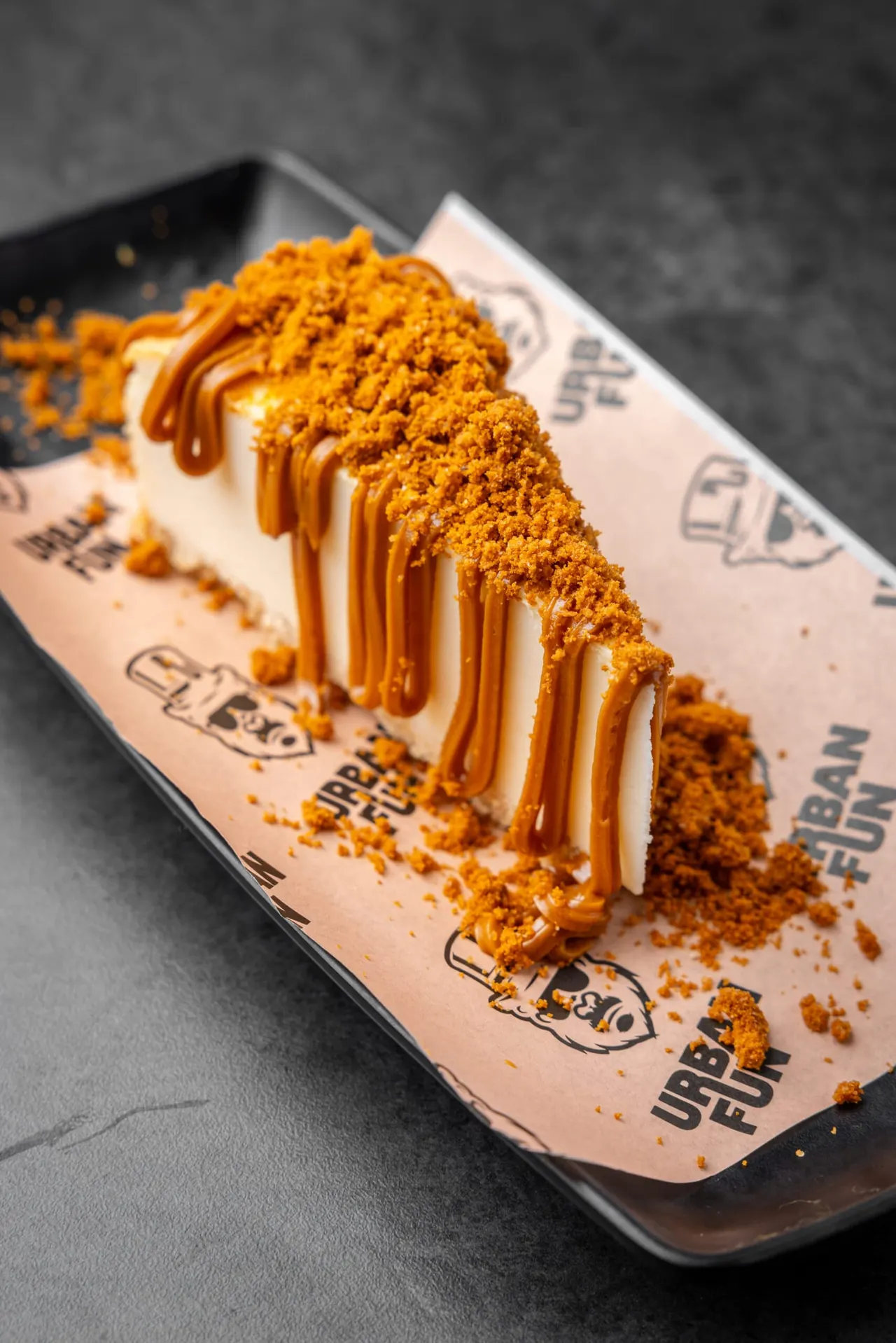 Dessert slice with caramel drizzle and biscuit crumb topping on a branded plate.