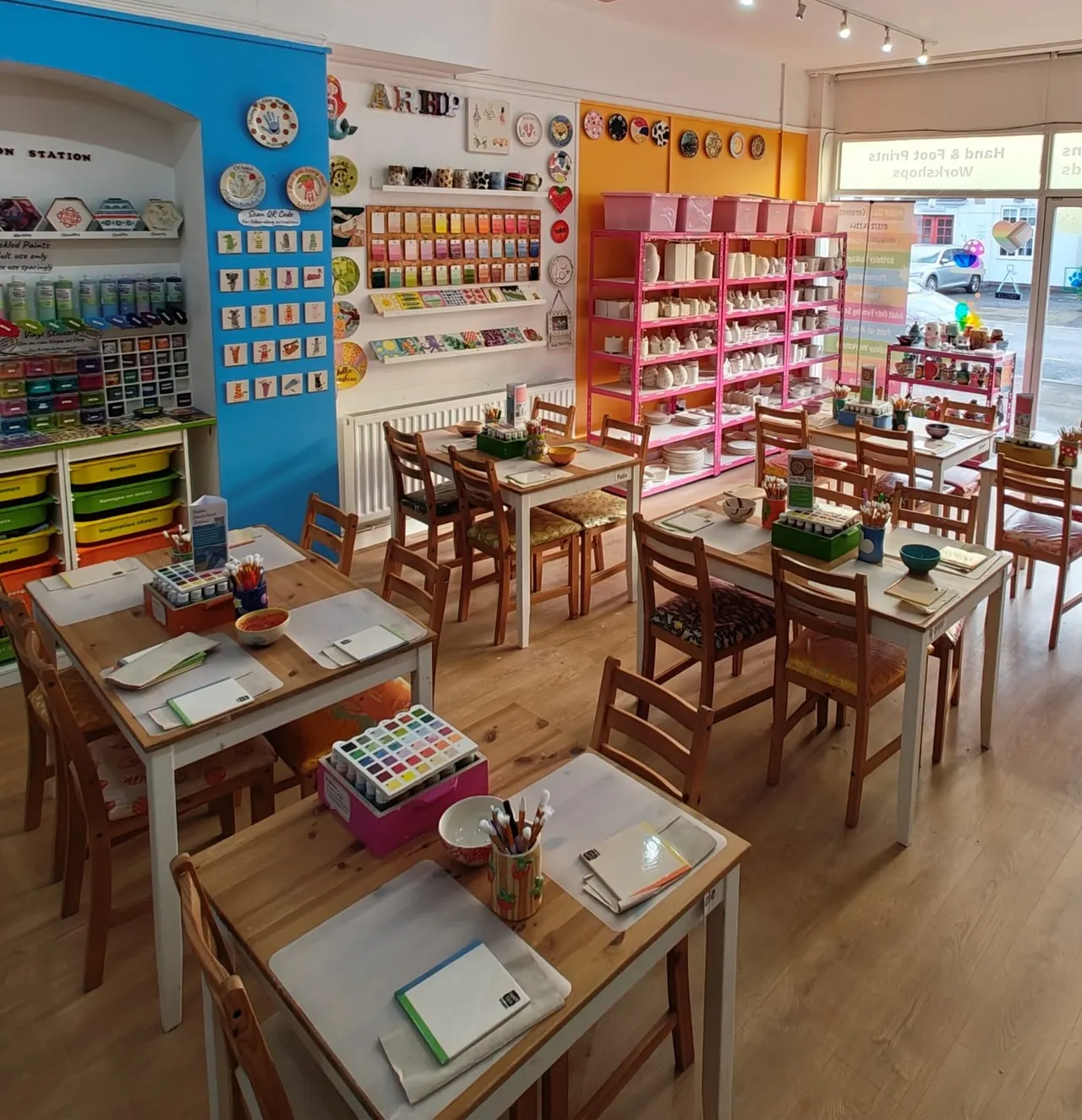 An art and crafts studio with wooden tables and chairs set up for pottery painting, surrounded by shelves of glazes, paints and ceramic supplies.