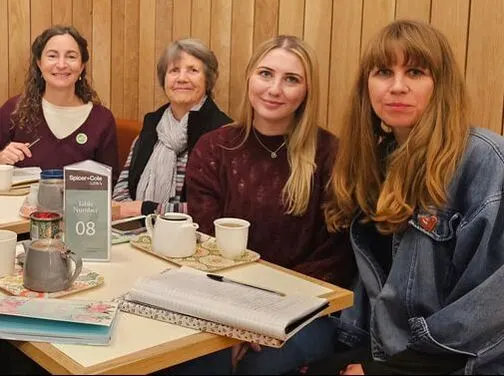 Four women of varying ages sit at a wooden table during a creative writing course, with tea, notebooks and course materials in front of them.