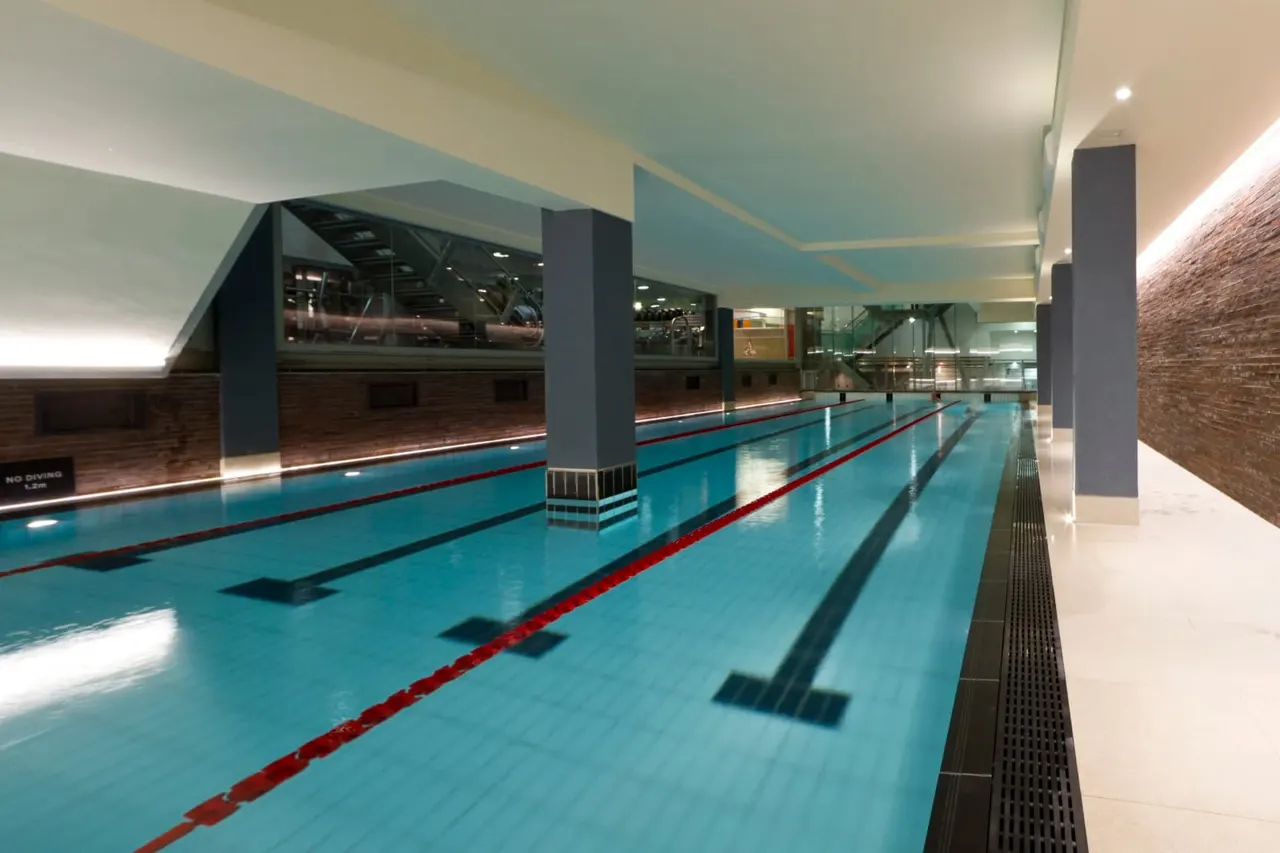 Empty indoor swimming pool with multiple lanes divided by red and white lane ropes, modern ceiling with blue panels, and dark support columns.