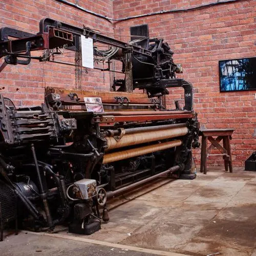 Antique textile loom machinery displayed against a red brick wall inside a gallery space.