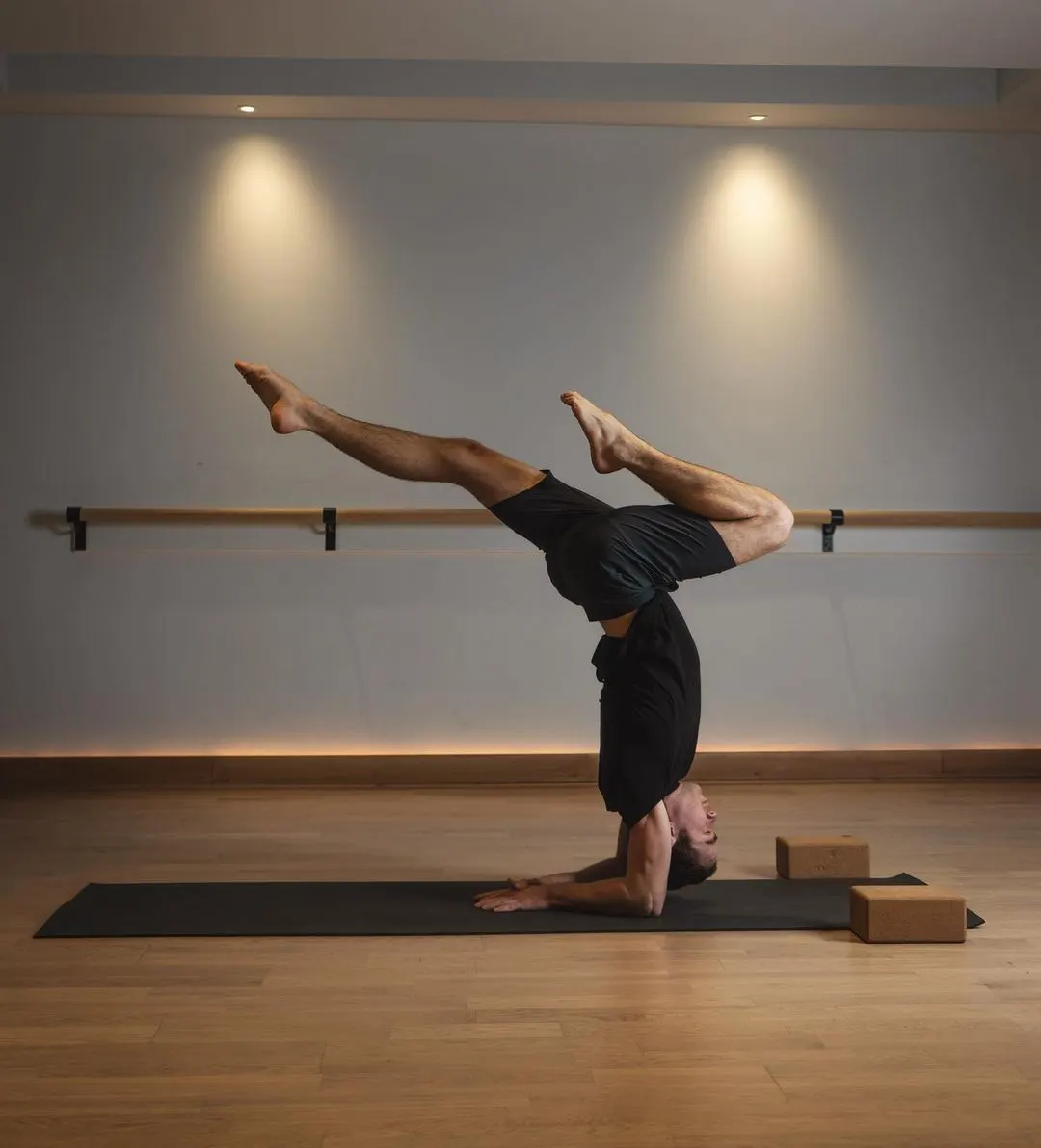 Man performing a headstand yoga pose on a mat in a studio with wooden flooring and ballet barre.
