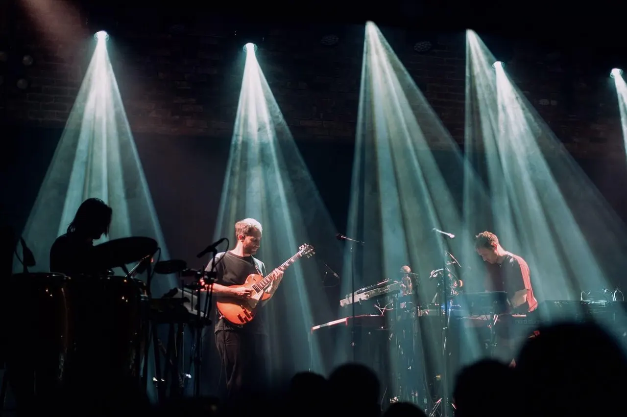 Band performing on stage with dramatic spotlight beams, guitarists and keyboardist playing to an audience.