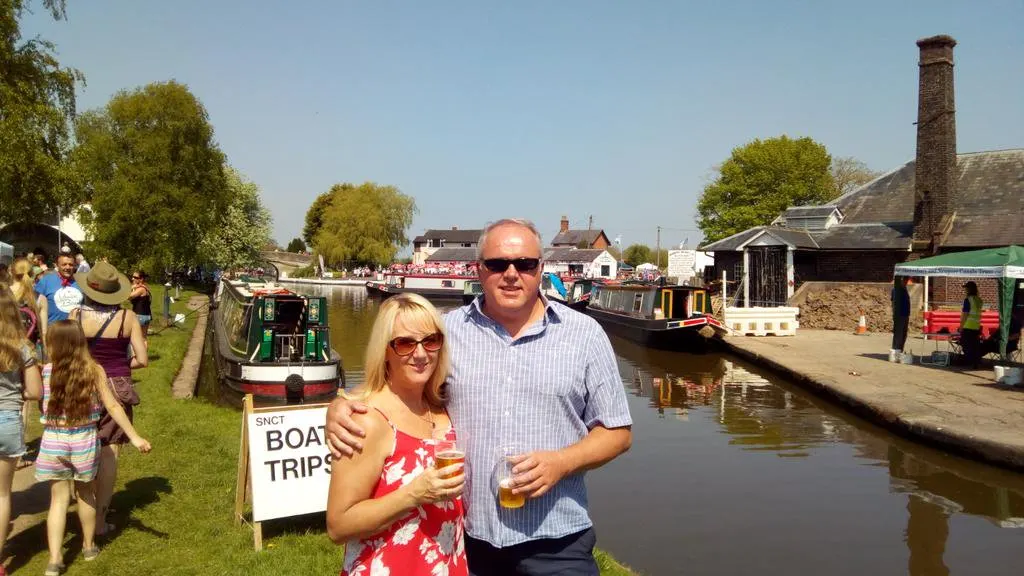 Couple holding drinks at a canal-side venue with narrowboats moored alongside, surrounded by visitors on a sunny day.